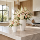 Two white textured ceramic vases filled with a large floral arrangement of white and blush pink roses, hydrangeas, and green foliage, displayed on a light marble kitchen island.