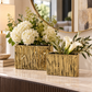 Two rectangular gold ceramic vases with a textured, tree-bark-like vertical pattern, filled with white flowers and green foliage, displayed on a marble console table in a modern interior setting.
