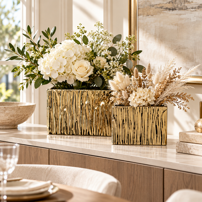 Two rectangular gold ceramic vases with a textured vertical bark-like pattern, filled with white and beige dried/germinal floral arrangements, displayed on a light marble console table in a bright, modern dining room.