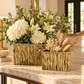 Two rectangular gold ceramic vases with a textured, metallic bark-like pattern, filled with white hydrangeas, lilies, and dried pampas grass, displayed on a polished marble console table in an elegant interior setting.