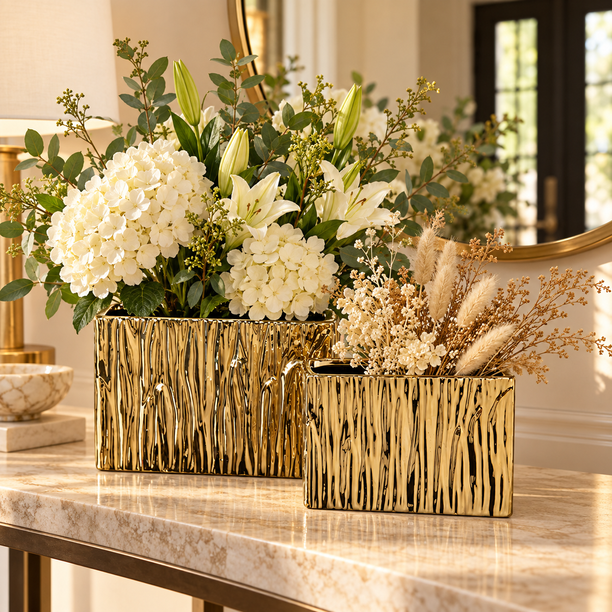 Two rectangular gold ceramic vases with a textured, metallic bark-like pattern, filled with white hydrangeas, lilies, and dried pampas grass, displayed on a polished marble console table in an elegant interior setting.