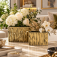 Two rectangular gold metallic vases with a textured, vertical wood-grain-like finish, filled with white and cream floral arrangements, displayed on a marble coffee table in a modern living room setting.