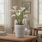 A decorative gray ceramic vase with a textured geometric pattern, filled with white calla lilies and green stems, placed on a wooden table in a softly lit living room.