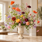 A large, vibrant wildflower arrangement in a distressed white stone urn vase, displayed on a marble countertop in a bright, modern living room.