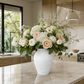 A large white textured ceramic vase filled with a lush floral arrangement of white and blush pink flowers with green foliage, set on a marble kitchen island.