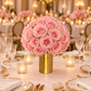 A large round bouquet of light pink roses arranged in a textured gold cylindrical vase on a decorated dining table with lit candles, crystal glasses, and gold tableware.