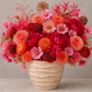 A vibrant autumnal floral arrangement featuring pink, red, and orange flowers in a textured beige ceramic pot, set against a neutral gray background.