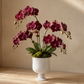 A potted artificial orchid arrangement with deep burgundy flowers, green leaves, and moss in a white pedestal vase, placed on a light wood surface against a beige wall.