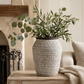 A gray and white patterned ceramic planter vase filled with green eucalyptus branches, styled on a wooden console table in a cozy living room setting next to a fireplace and sofa.