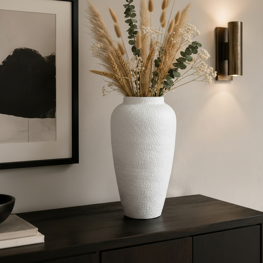 A tall textured white ceramic vase filled with dried wheat, pampas grass, and green eucalyptus stems, displayed on a dark wood console table in a modern interior setting.