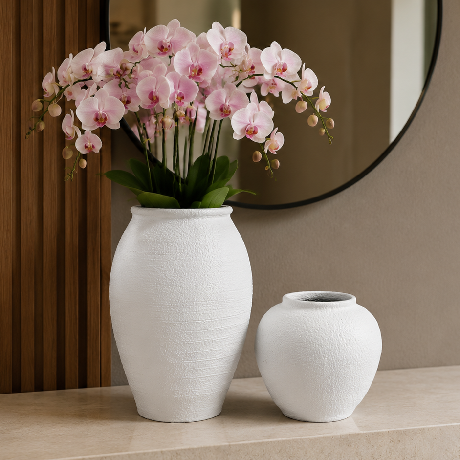 Two white textured ceramic vases, one tall with pink orchids and one short, displayed on a light stone console table in a modern interior setting.