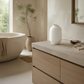 A white textured ceramic vase displayed on a light stone vanity in a softly lit bathroom setting with a bathtub, potted plant, and mirror.