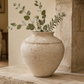 A large textured cream-colored stone urn vase filled with green eucalyptus branches, displayed on a stone mantel in a cozy living room setting.