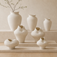 A set of seven organic-shaped ceramic vases in a warm off-white, textured stoneware finish, displayed on minimalist stone risers in a modern, sunlit room with beige walls and wood flooring.