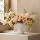 A white ribbed ceramic urn vase filled with a soft blush and cream floral arrangement, displayed on a stone pedestal in a warm, sunlit interior setting.
