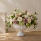 A large white ribbed urn-shaped vase filled with a lush floral arrangement of cream, blush, and lavender roses, green hydrangeas, and trailing greenery, displayed on a wooden table against a textured beige wall.