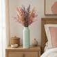 A tall light aqua ceramic vase filled with a dried floral arrangement in pink, peach, and lavender tones, styled on a wooden bedside table next to a bed with a pink pillow.