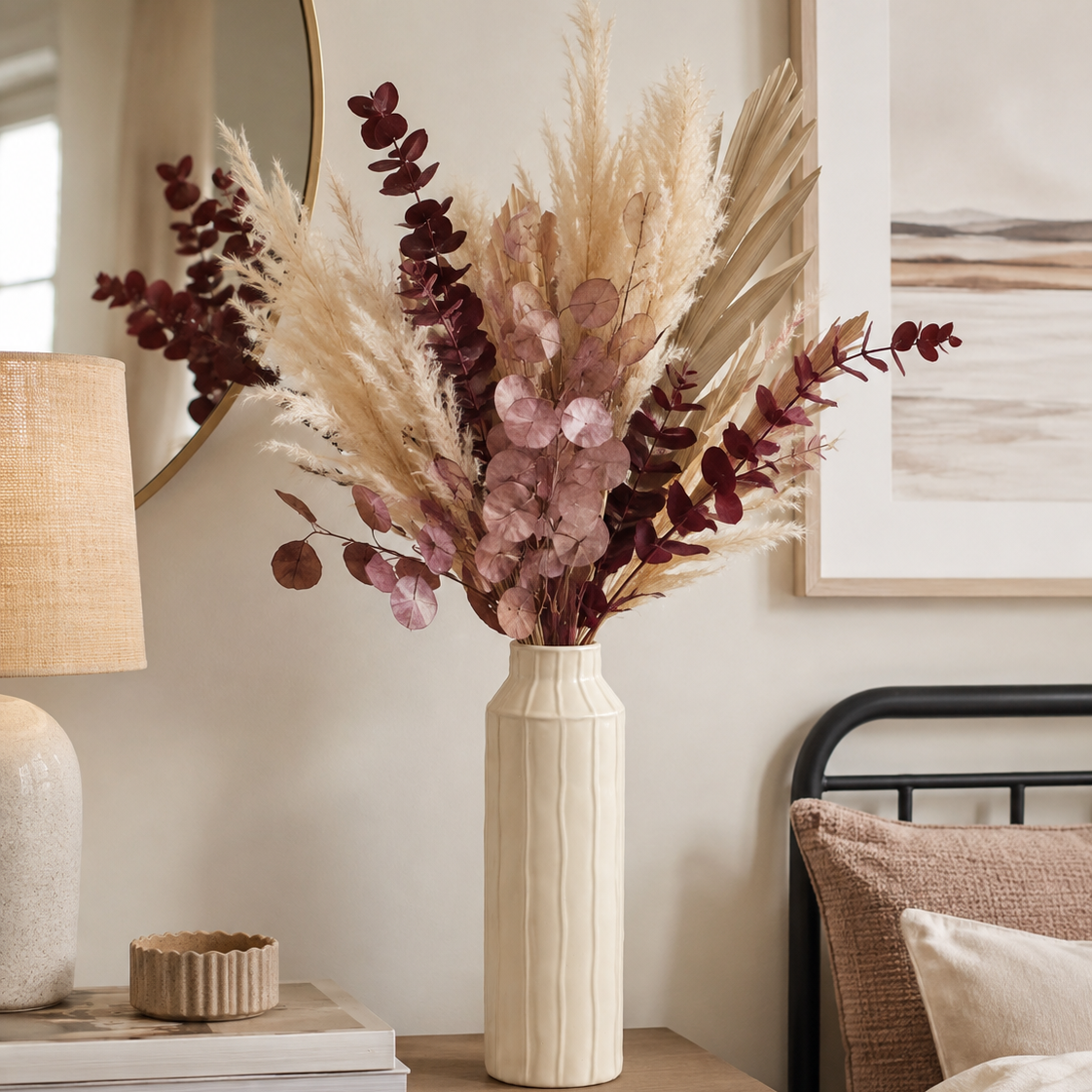 A dried floral arrangement in a ribbed cream ceramic vase, styled on a wooden bedside table in a bedroom setting next to a lamp and bed.