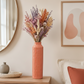 A tall ribbed terracotta pink ceramic vase filled with a colorful assortment of dried flowers, displayed on a light wood side table in a cozy, modern room setting.