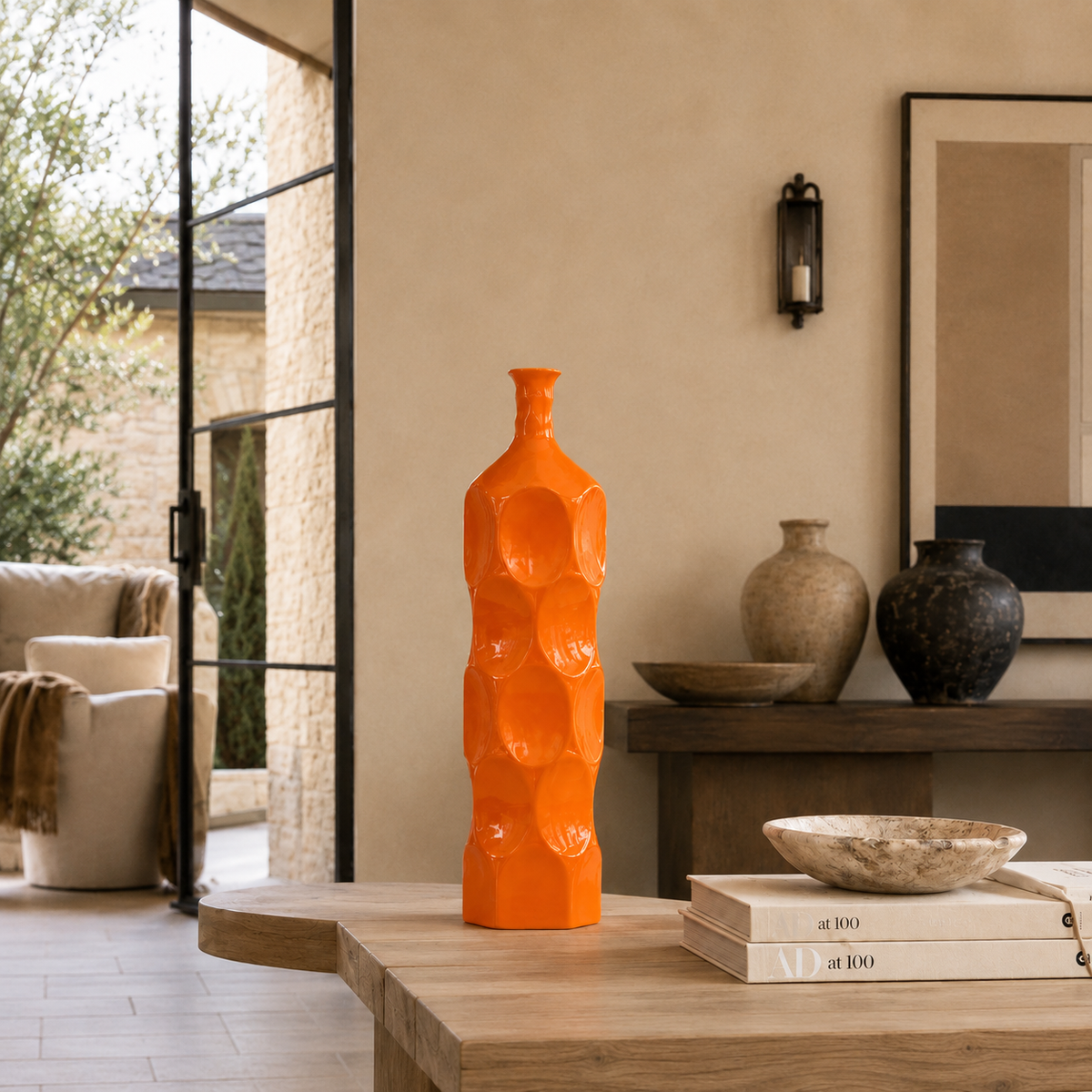 A tall, bright orange sculptural ceramic vase with a textured, bubble-like pattern stands on a light wood table in a modern, warm-toned living room setting.
