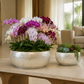 Two silver planters displayed on a light stone table in a modern living room. The larger pot holds a vibrant arrangement of purple, pink, and white orchids, while the smaller pot contains green succulents.
