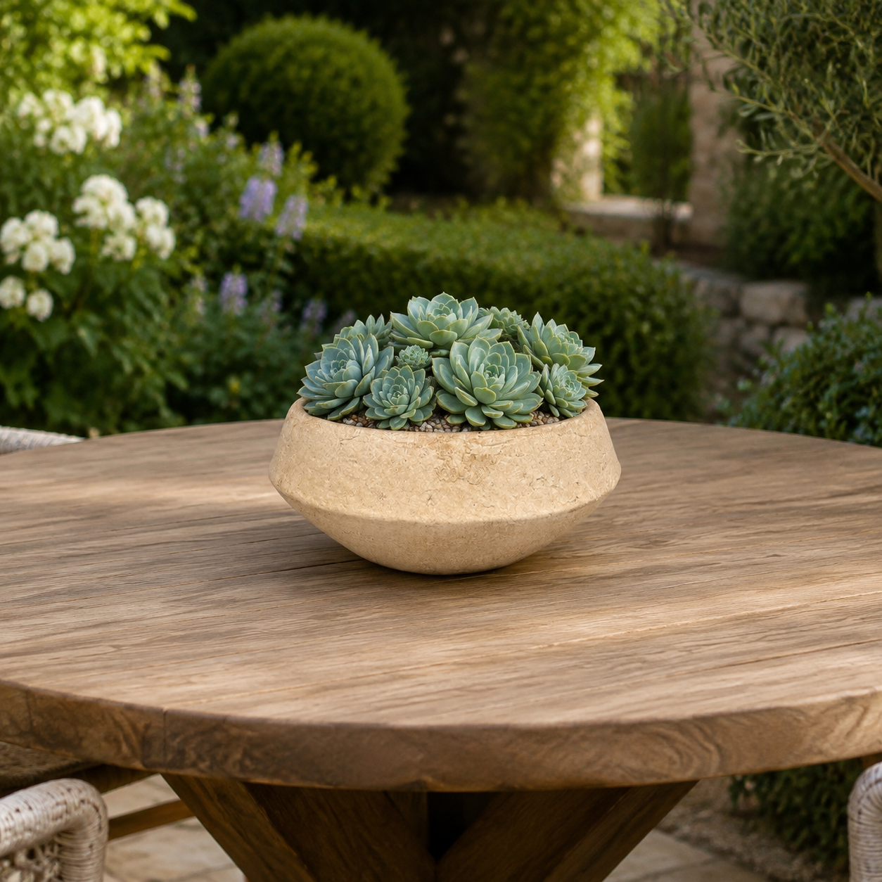 A round beige stone planter filled with green succulent plants, set on a round wooden outdoor table with a blurred garden background.