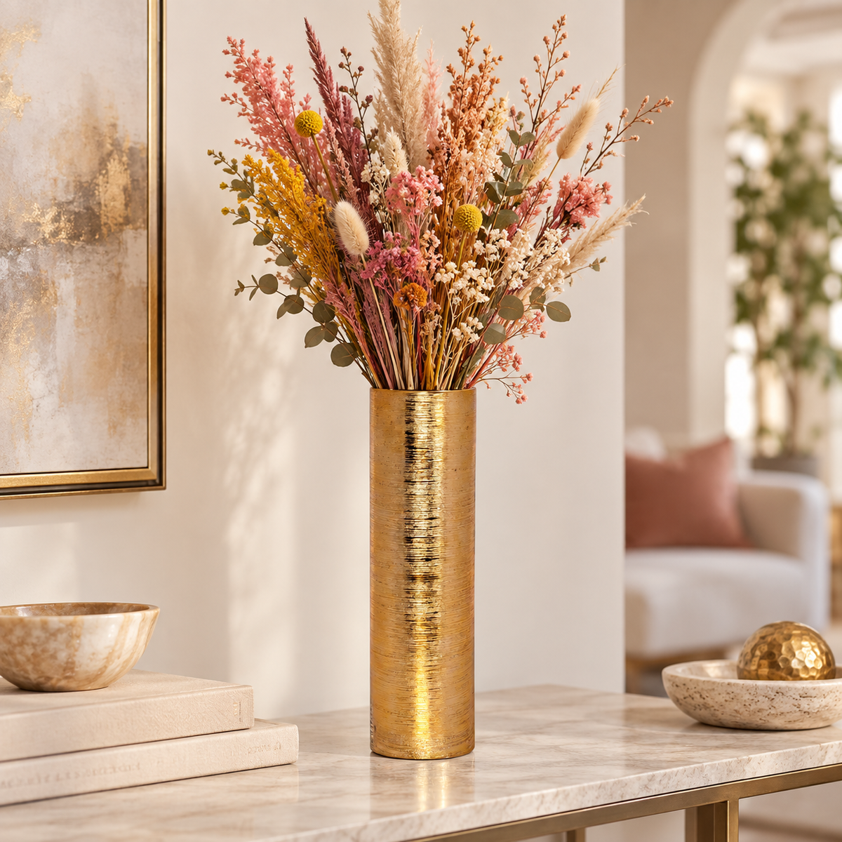 A tall cylindrical gold vase filled with a colorful assortment of dried flowers and grasses, displayed on a marble console table in a modern, softly lit living room.