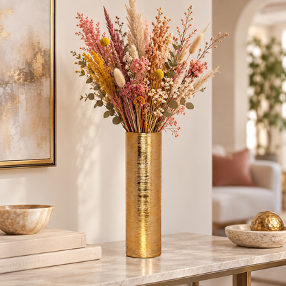 A tall cylindrical gold vase filled with a colorful assortment of dried flowers and grasses, displayed on a marble console table in a modern, softly lit living room.