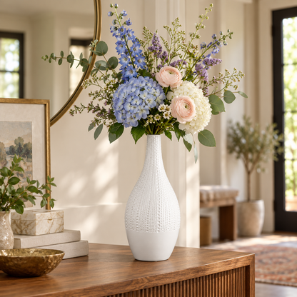 A white textured ceramic vase filled with a floral arrangement of blue hydrangeas, blush pink roses, white peonies, and green foliage, displayed on a wooden console table in a bright, modern living room.