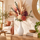 A dried floral arrangement in a textured white ceramic vase, styled on a wooden console table in a warm, sunlit living room.