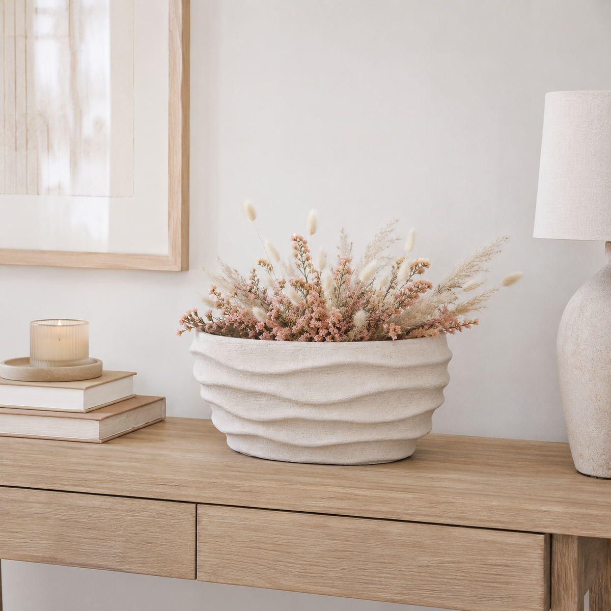 A textured cream-colored ceramic planter with wavy horizontal ridges, filled with dried pink and beige flowers, styled on a light wood console table next to a candle, books, and a lamp.