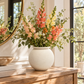 A ceramic white vase filled with a colorful floral arrangement of pink, orange, and yellow flowers with green foliage, displayed on a wooden console table in a bright room.