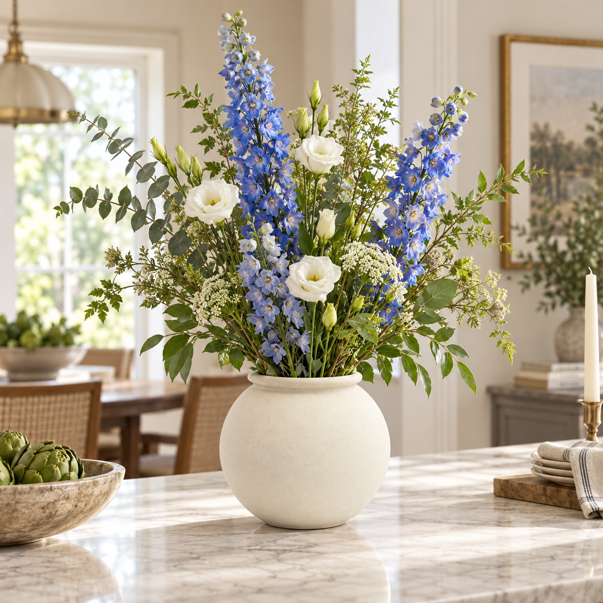 A floral arrangement in a round cream ceramic vase on a marble countertop, featuring blue delphiniums, white roses, and green foliage in a bright dining room setting.