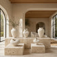 A set of sculptural, organic-shaped off-white ceramic vases displayed on stone plinths inside a modern, sunlit room with beige walls and large windows.