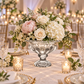 A floral centerpiece arrangement in a clear ornate pedestal bowl, set on a decorated dining table with lit candles, crystal glasses, and warm bokeh lights in the background.