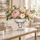 A clear glass pedestal bowl filled with a floral arrangement of pink and cream roses, ranunculus, and green eucalyptus, displayed on a marble and gold console table in a bright, elegant living room.