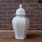A white ceramic lidded jar with a pointed top, standing on a wooden surface against a red brick wall background.