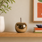 A shiny metallic gold apple-shaped decorative object with a short stem sits on a light wood shelf in a styled interior setting, next to a white ribbed vase with green leaves and a stack of books with a small purple bowl.