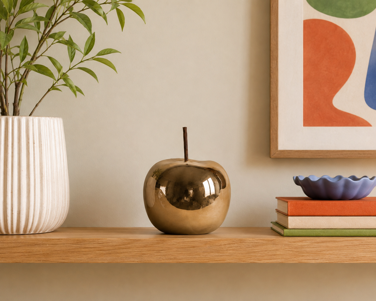 A shiny metallic gold apple-shaped decorative object with a short stem sits on a light wood shelf in a styled interior setting, next to a white ribbed vase with green leaves and a stack of books with a small purple bowl.