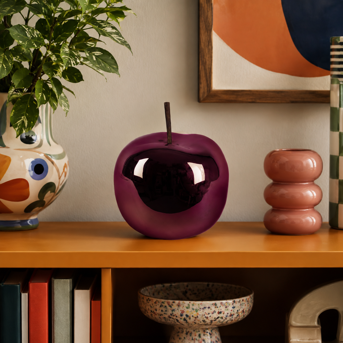 A glossy purple apple-shaped decorative object with a dark reflective center sits on a wooden shelf in a styled interior setting with books, a plant, and abstract wall art.