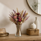 A clear glass vase filled with a dried floral arrangement in muted pink, burgundy, beige, and purple tones, styled on a wooden console table in a warm interior setting.