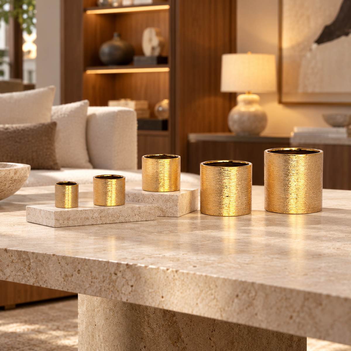 Five textured gold cylindrical candle holders of varying sizes arranged on matching rectangular stone risers on a light beige travertine coffee table in a modern, warm-toned living room.