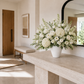A large white ceramic vase filled with a lush floral arrangement of white roses, hydrangeas, and green foliage, styled on a light stone console table in a bright, modern entryway.