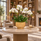 A potted white Phalaenopsis orchid plant with green leaves in a textured gold ceramic pot, displayed on a round marble-top table in a luxurious, warm-toned study with bookshelves and a fireplace.