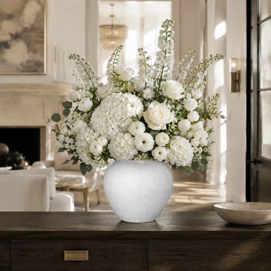 A large white ceramic vase filled with a lush white floral arrangement featuring hydrangeas, roses, ranunculus, and green foliage, displayed on a dark wood console table in a bright, elegant living room.