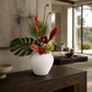 A white textured ceramic vase filled with a tropical floral arrangement, including green monstera leaves, orange bird of paradise, red anthurums, and green orchids, displayed on a dark wooden console table in a modern minimalist living room.