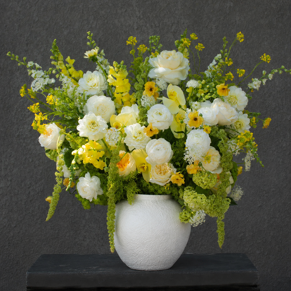 A large floral arrangement featuring white and yellow flowers with green foliage, presented in a textured white ceramic vase on a dark surface against a dark gray wall.