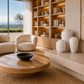 A modern living room interior featuring a round wooden coffee table, a beige upholstered armchair, a matching sofa partial view, a built-in wooden bookshelf with warm lighting, and several large white ceramic vases on a stone ledge.